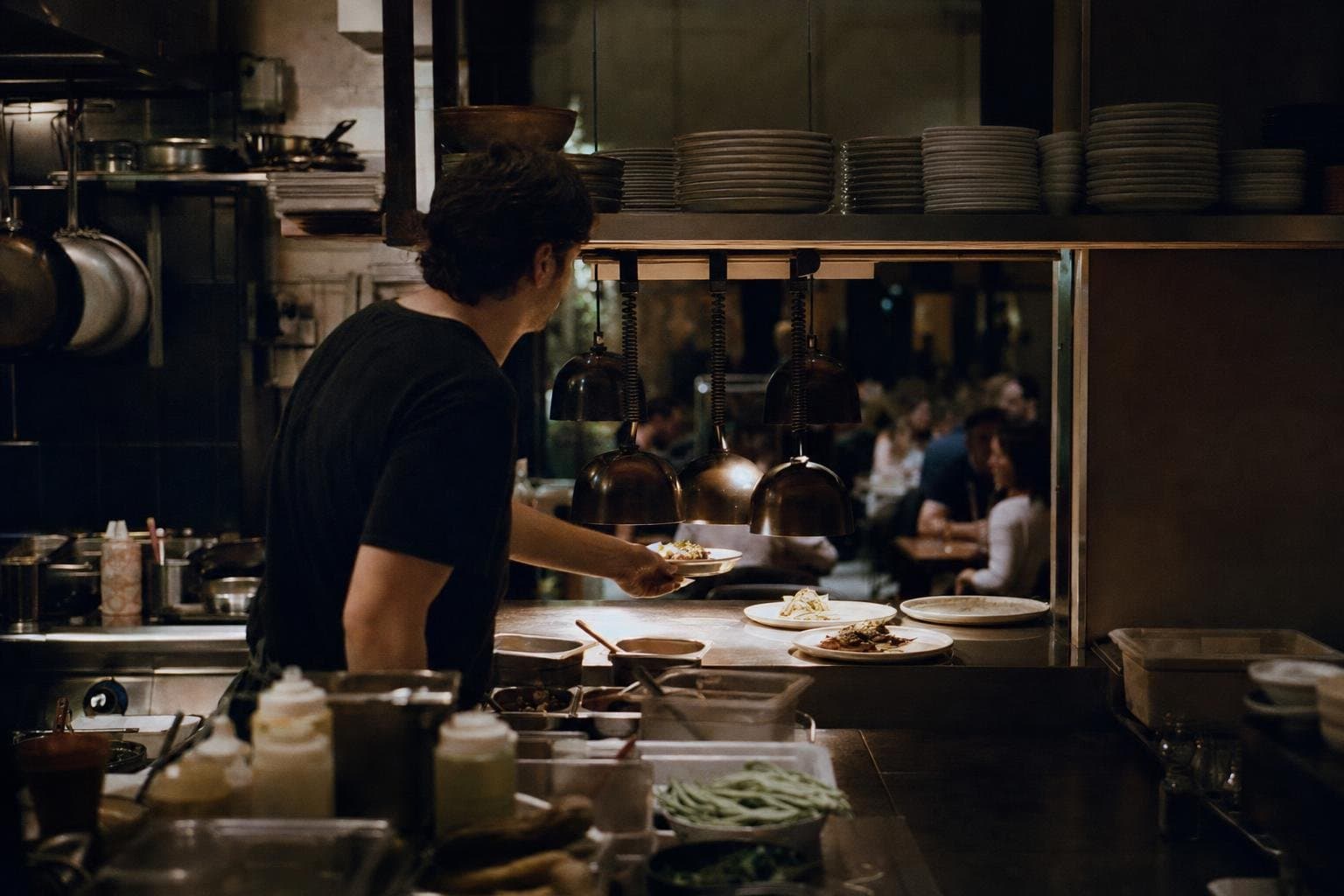 The kitchen pass of a busy independent restaurant during evening service, finished plates waiting under heat lamps