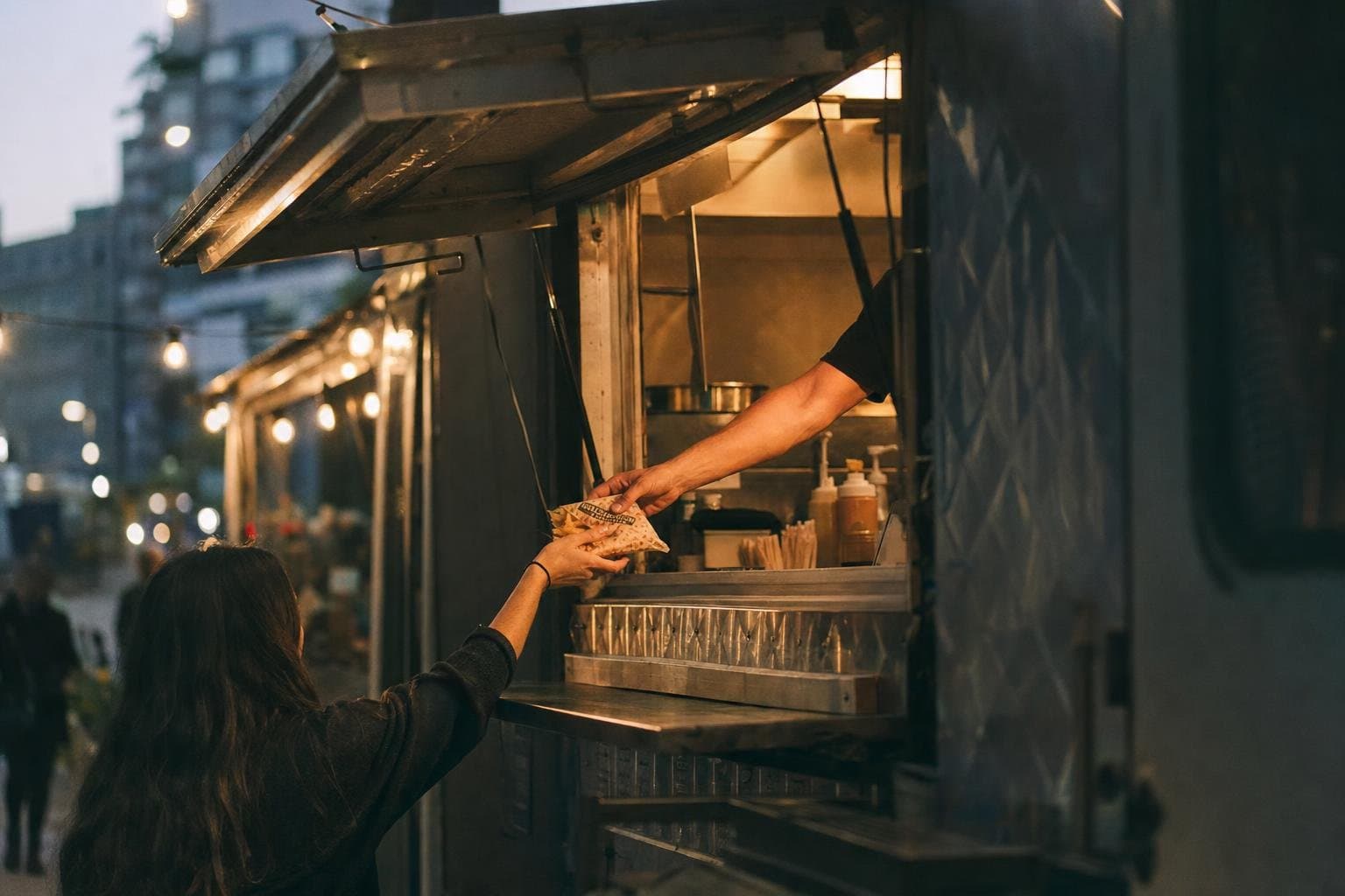 The glowing service window of a food truck at dusk with a hand passing food out